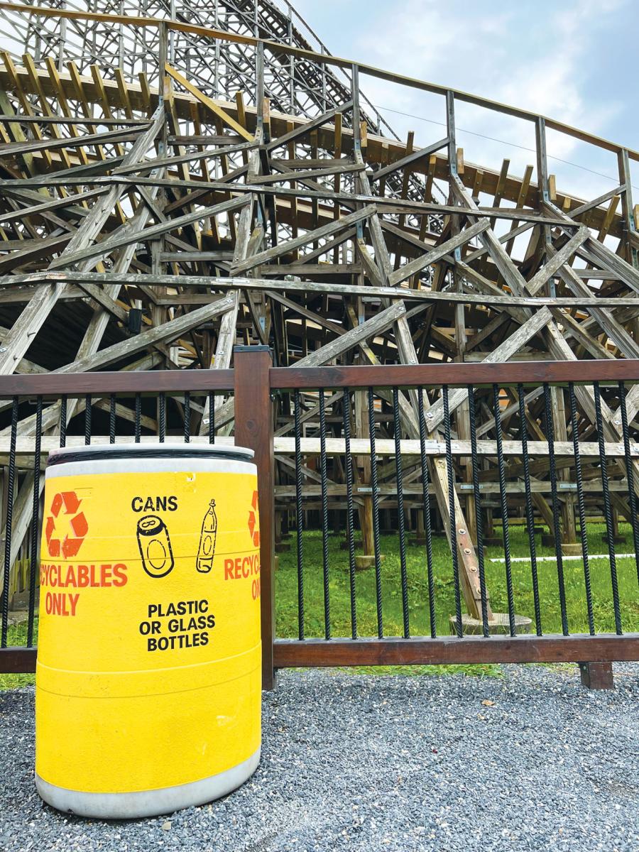 A yellow trash bin forward-facing a Knoebels Amusement Resort roller coaster, indicating "Plastic or Glass Bottles" recycling instructions