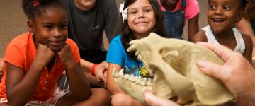Children are gathered in a circle, observing a skeletal mock-up of a dinosaur's head