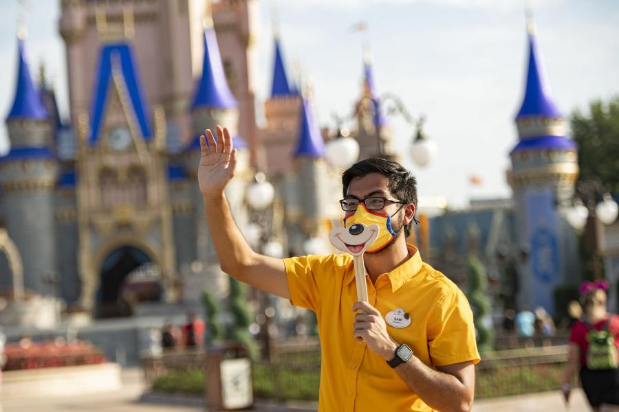Un employé de Magic Kingdom salue les invités tout en portant un masque et tenant un bâton avec une photo du sourire de Mickey imprimée dessus.