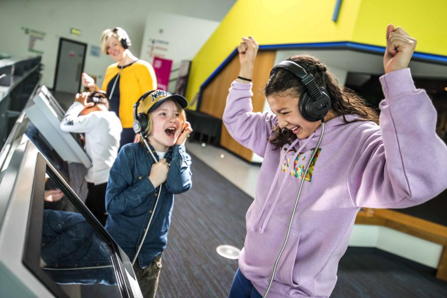 Two children cheer at an exhibit while wearing headphones.