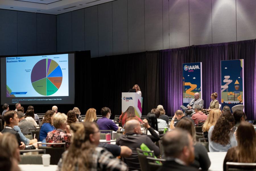 Wide-angle view of the "Finding Your Revenue" EDUSession, with attendees looking at a PowerPoint display on the left side featuring a pie chart calculating Denver Zoo's business model
