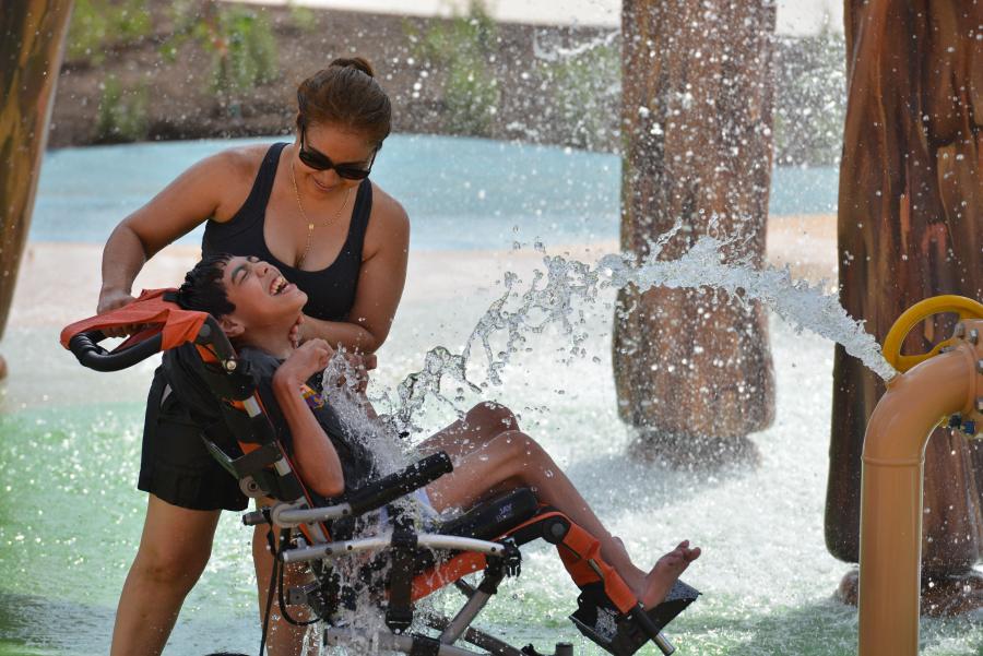 A parent and child with support needs enjoying water splash fountain inside Morgan’s Wonderland in San Antonio