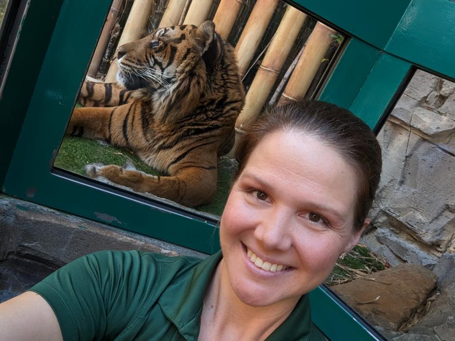 Kayla sonríe en una selfie con Bandar el tigre en su recinto al fondo.