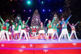 Artista en el escenario vistiendo trajes festivos y en el fondo central, la mascota de Snoopy disfrazada de Papá Noel también actuando en un espectáculo navideño en King's Dominion.