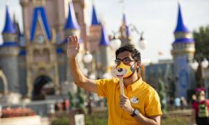 A Magic Kingdom employee waves at guests while wearing a mask and holding a stick with a picture of Mickey's smile printed on it.